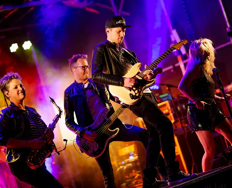 Band speelt live muziek op een podium met een zangeres, gitarist, bassist en saxofonist verlicht door kleurrijke lichten.