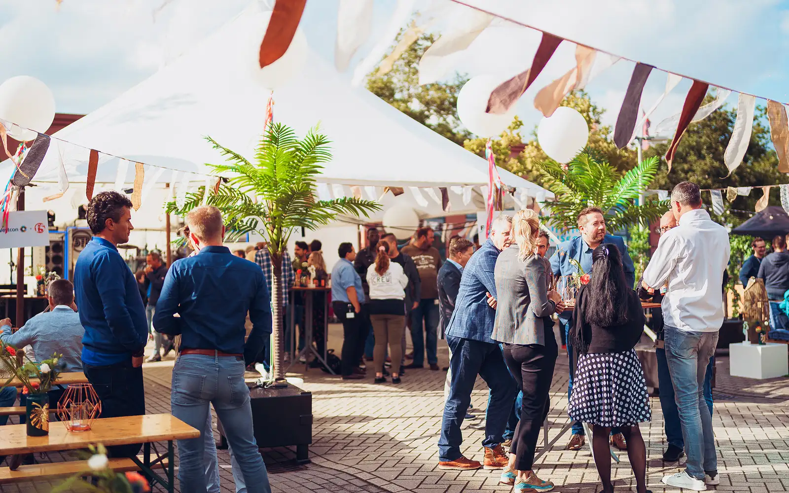Mensen praten en staan bijeen onder een feestelijke buiten tent met vlaggenlijnen en planten.