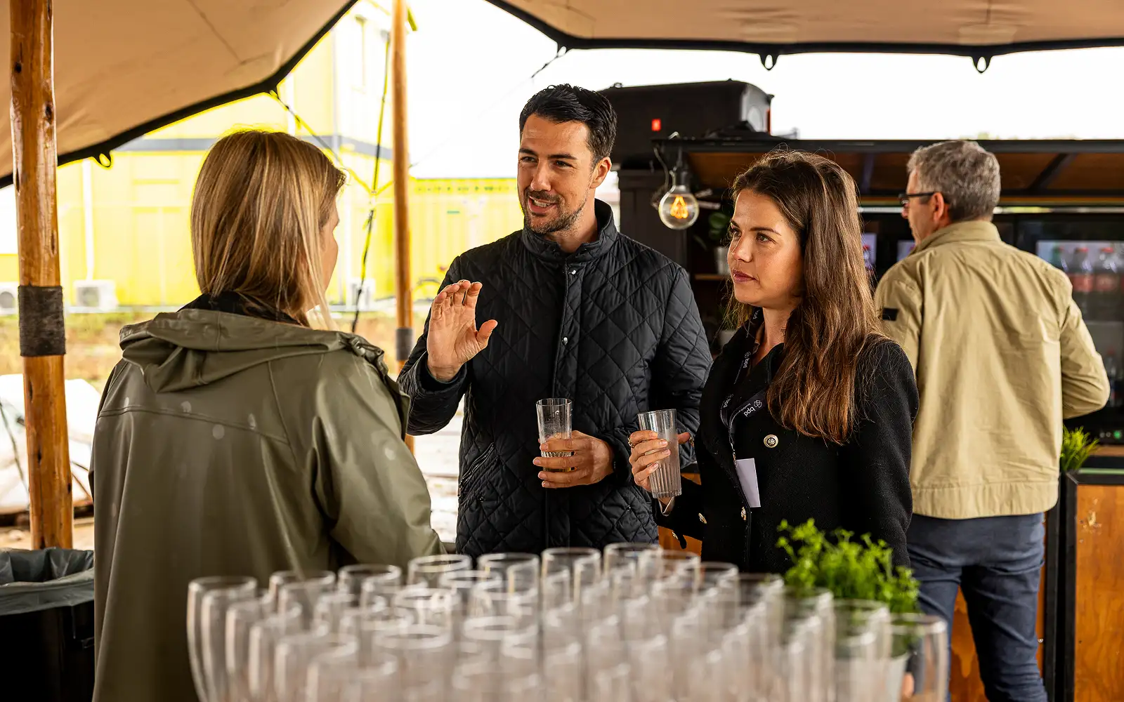 Drie volwassenen praten en houden glazen vast onder een tent met vele lege glazen op de voorgrond.