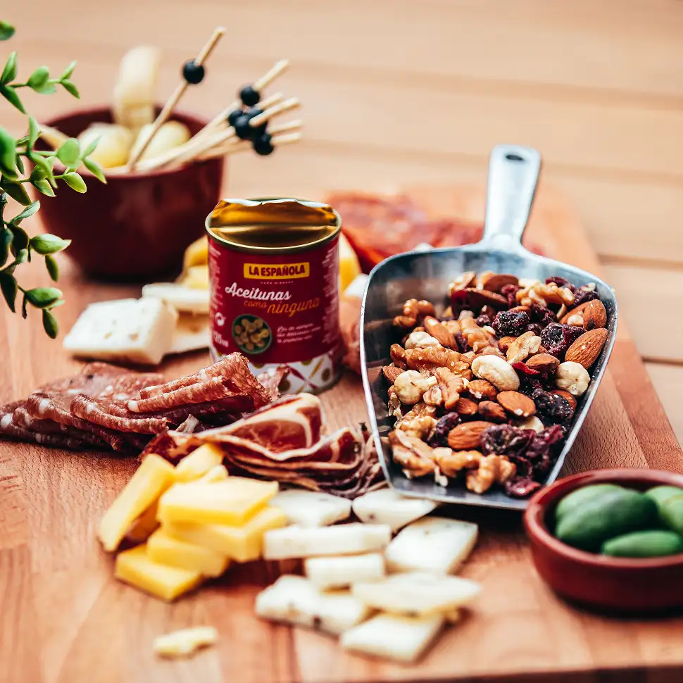 Hapjesplank met diverse kazen, vleeswaren, notenmix in een metalen schep en een open blikje olijven.