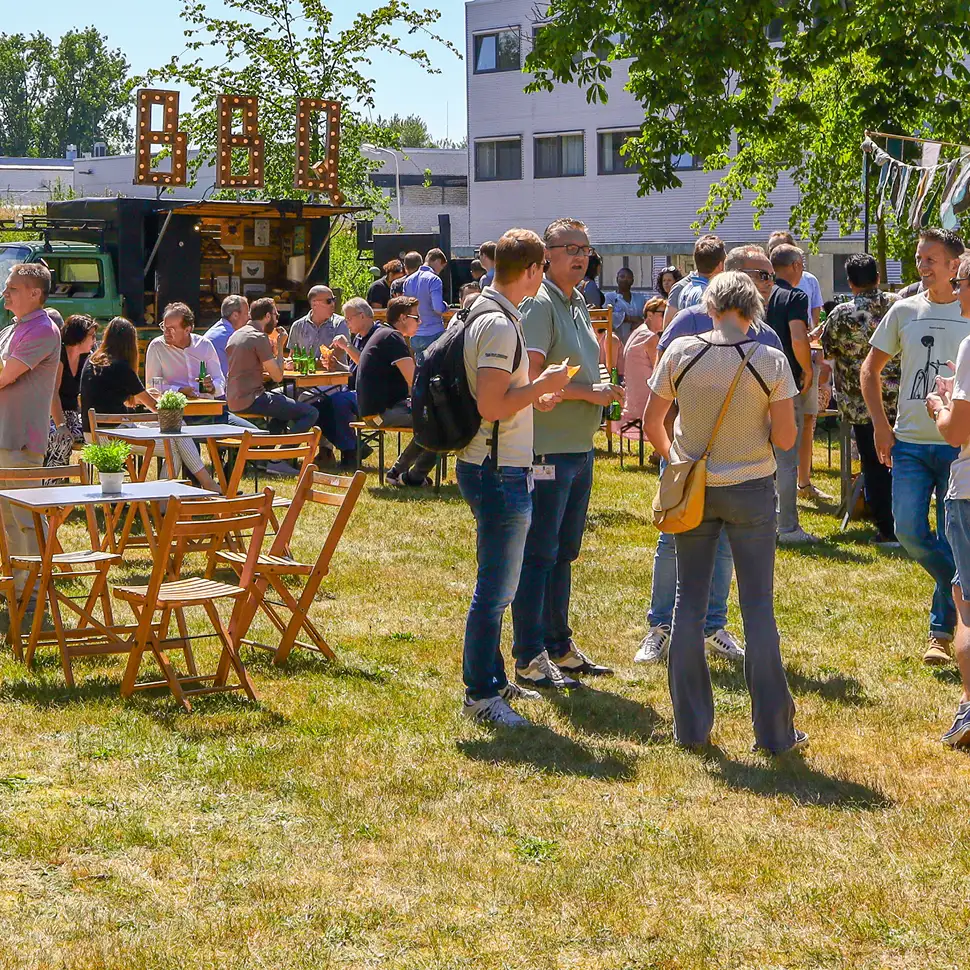 Mensen genieten van een BBQ-evenement buiten, met tafels en stoelen op gras en een foodtruck met verlichte letters 'BBQ'.
