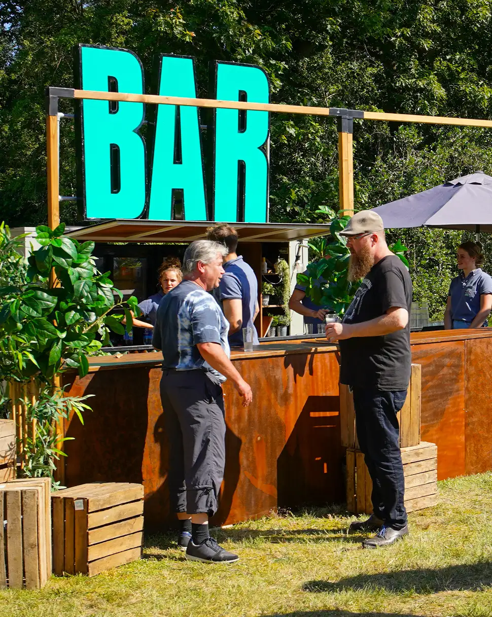 Twee mannen praten bij een bar in de buitenlucht onder een groot groen bord met de tekst 'BAR', omgeven door planten en houten kratten.