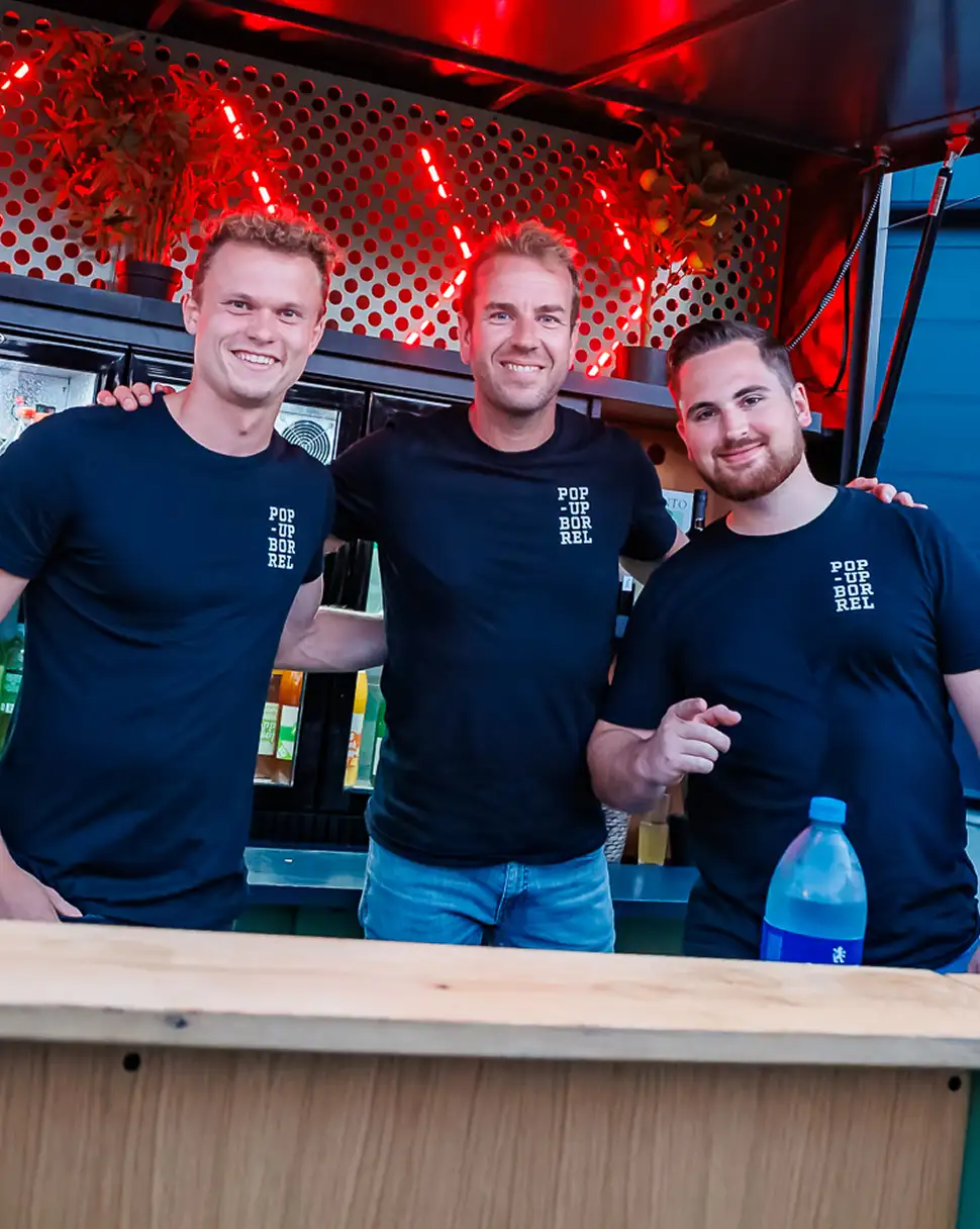 Drie lachende mannen in zwarte T-shirts met 'POP UP BORREL' achter een bar met rode verlichting.