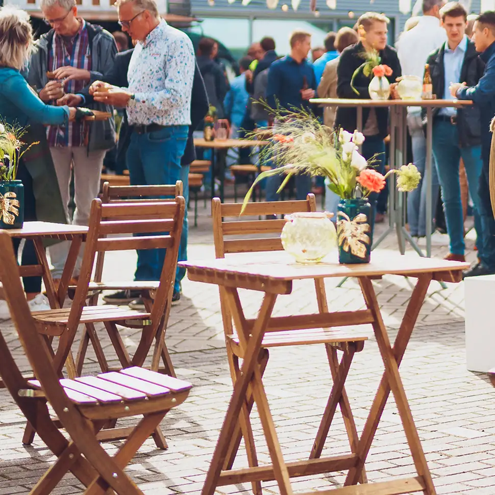 Houten klaptafels en stoelen met bloemstukken, mensen praten en staan rondom hoge tafels op een sfeervol buitenfeest.