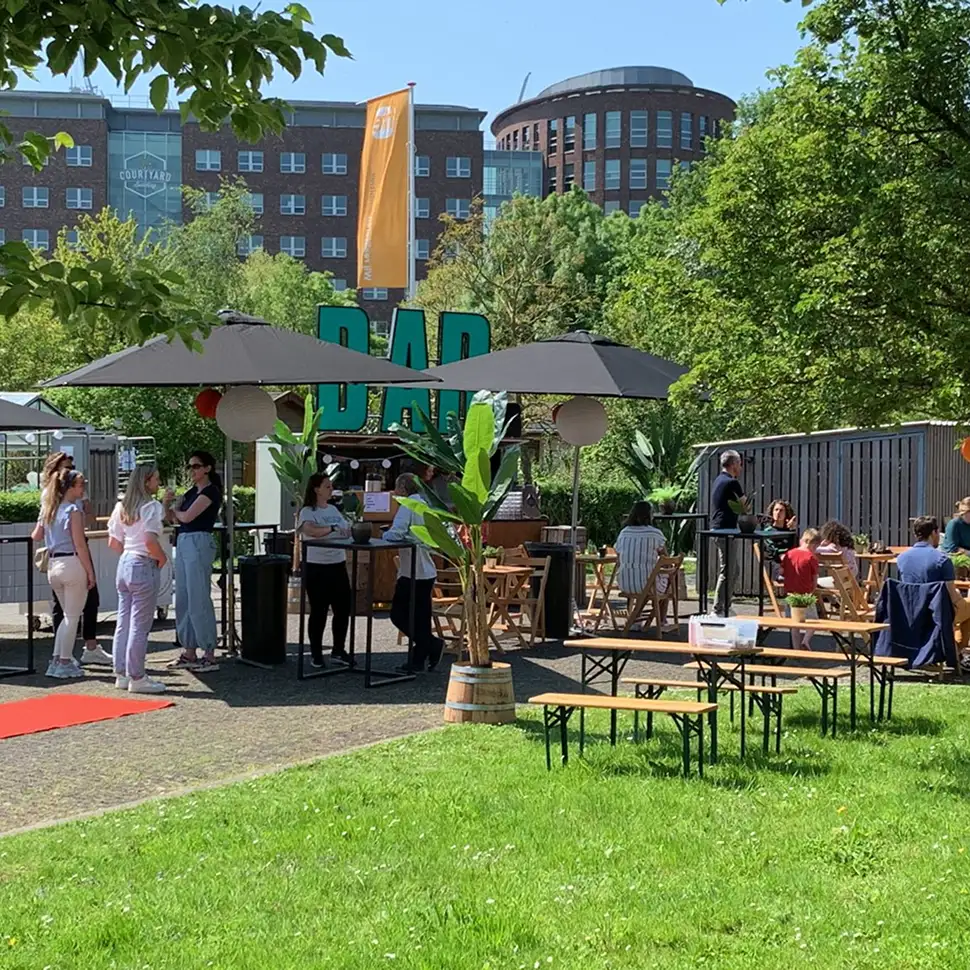 Buitenterras met mensen bij een bar onder grote parasols, omringd door groen en gebouwen op een zonnige dag.
