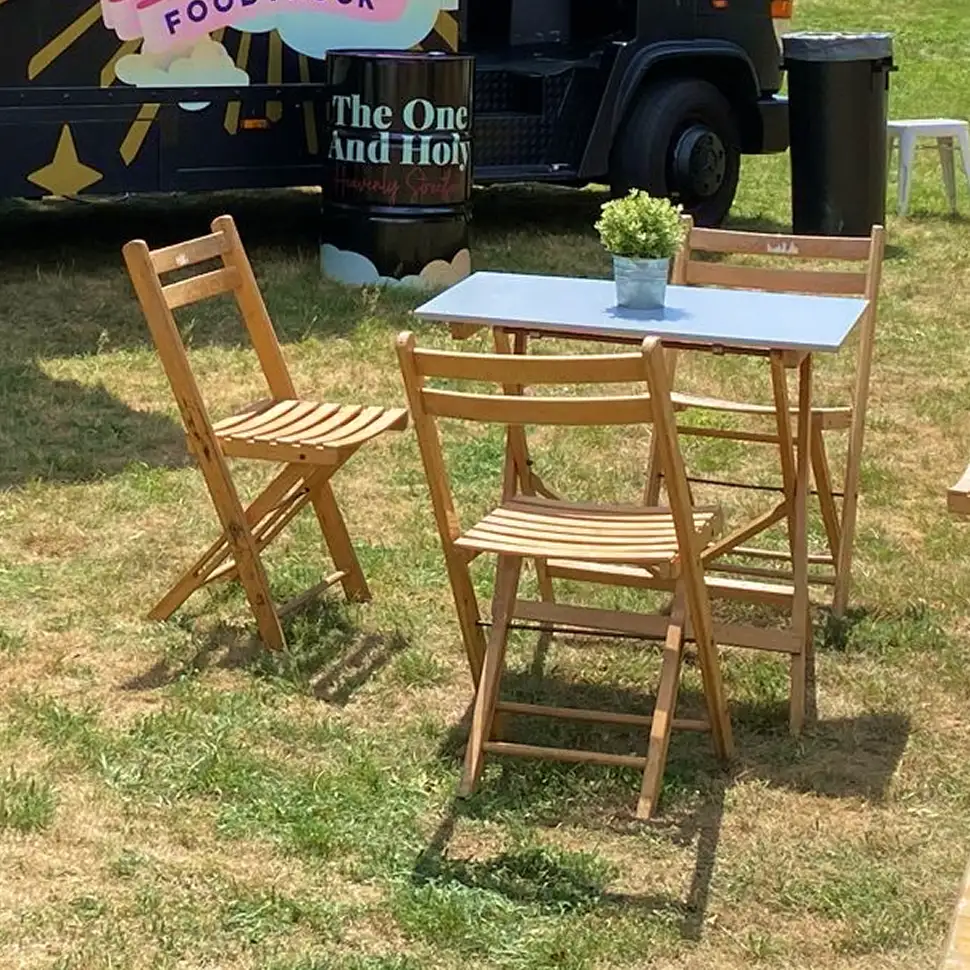 Houten tuintafel met een kleine plantenpot erop, omringd door drie houten klapstoelen op een grasveld.