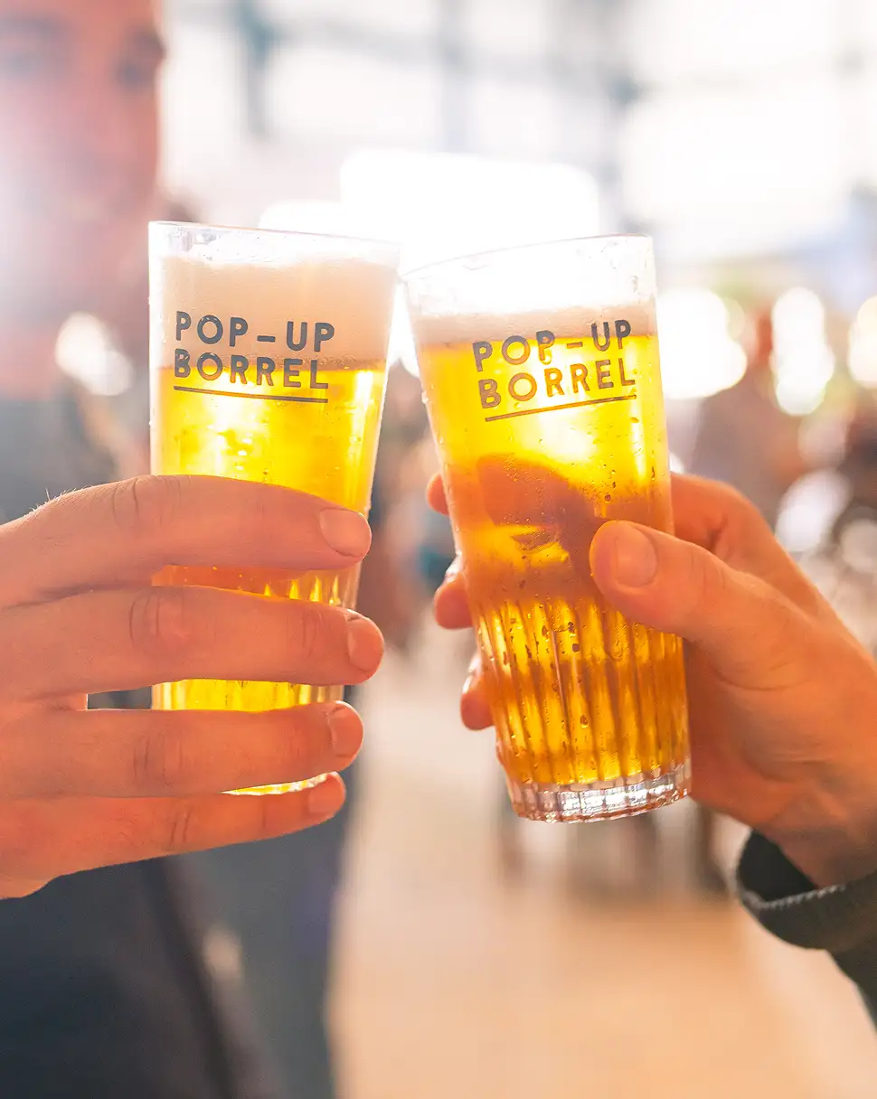 Twee mensen die biertjes proosten met glazen waarop 'Pop-up Borrel' staat.