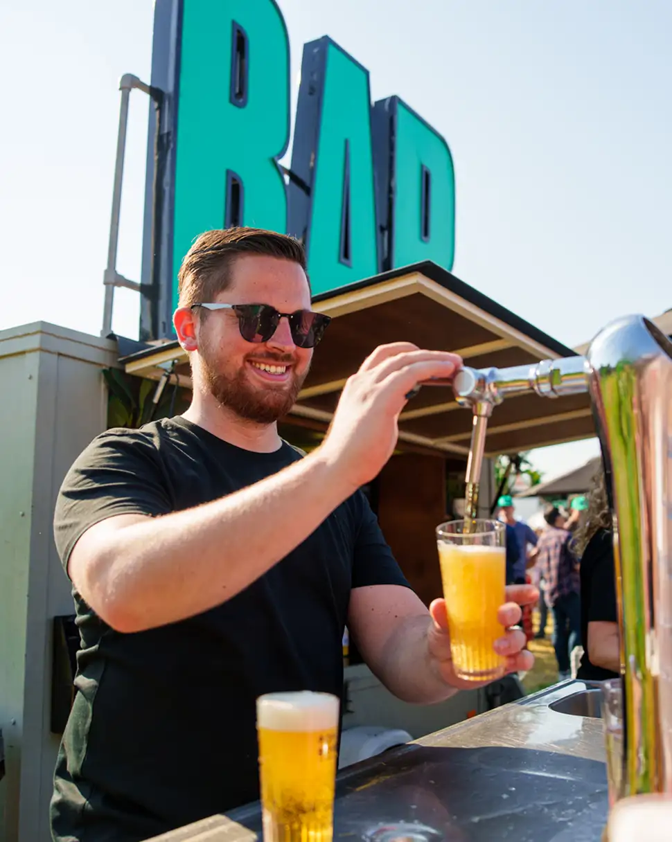 Man met zonnebril schenkt een glas bier in bij een buitenbar met groot groen bord 'BAR' op de achtergrond.