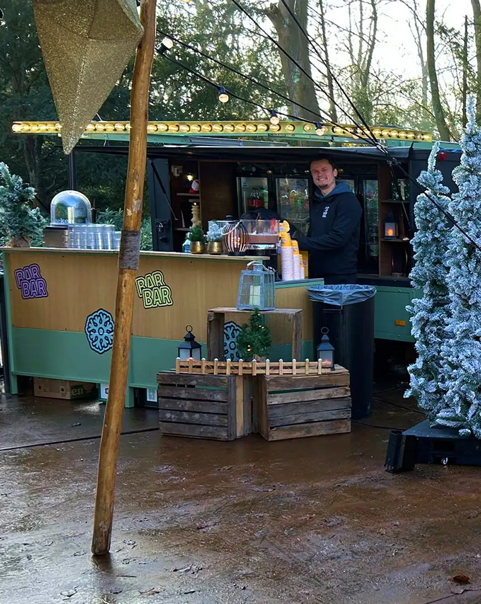 Bar met houten kisten en kerstdecoraties buiten, bediend door lachende man achter de counter.