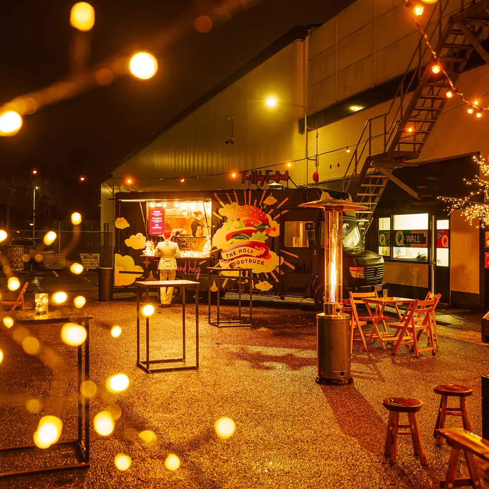 Sfeervolle nachtelijke foodtruck met warme verlichting, buiten tafels en stoelen op een natte ondergrond.