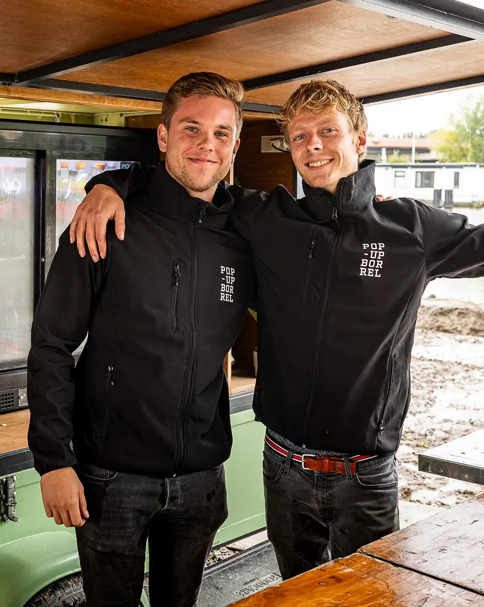 Twee jonge mannen in zwarte jassen met 'POP UP BORREL' tekst, lachend en met armen om elkaar heen.