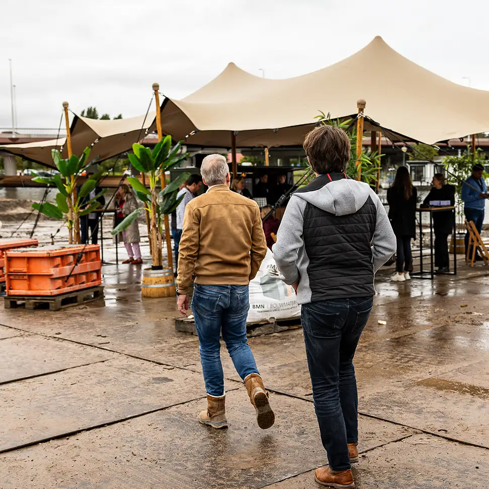Twee mannen lopen naar een marktkraam met tentdoeken en groene planten op een natte betonnen ondergrond.
