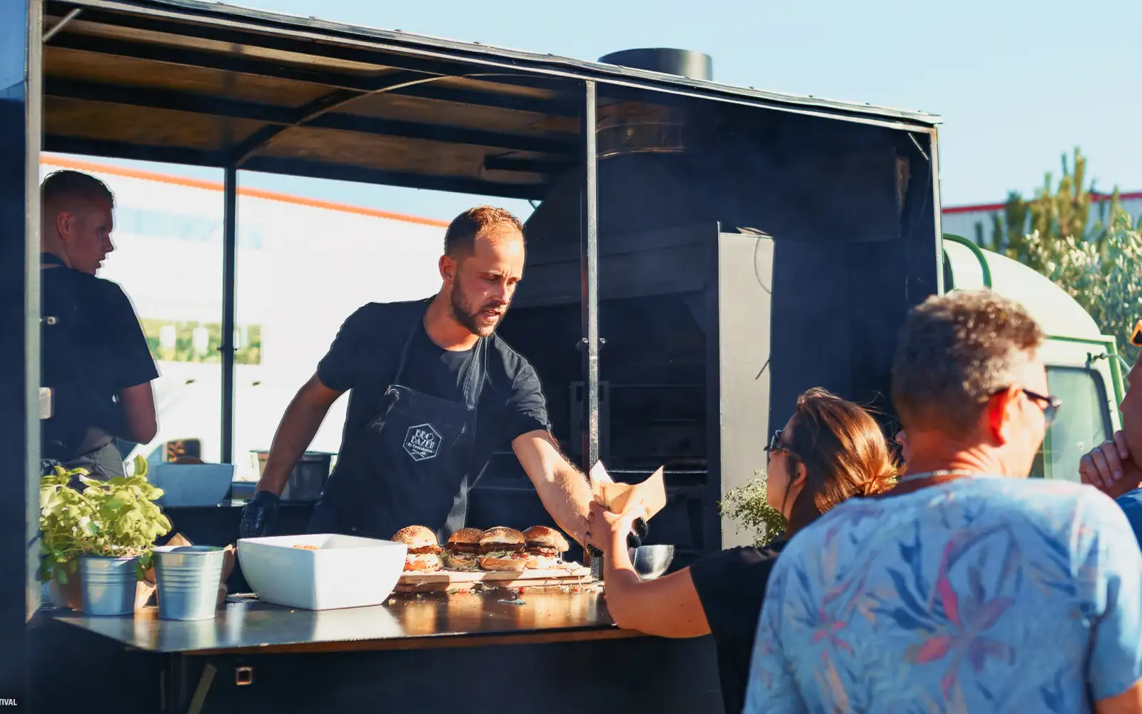 Man in een zwart schort serveert hamburgers vanuit een foodtruck aan een vrouw in de rij tijdens een zonnige dag.