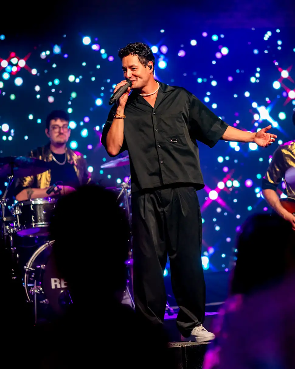 Zanger in zwart overhemd en broek staat op het podium met microfoon, achtergrond gevuld met kleurrijke lichtpunten.