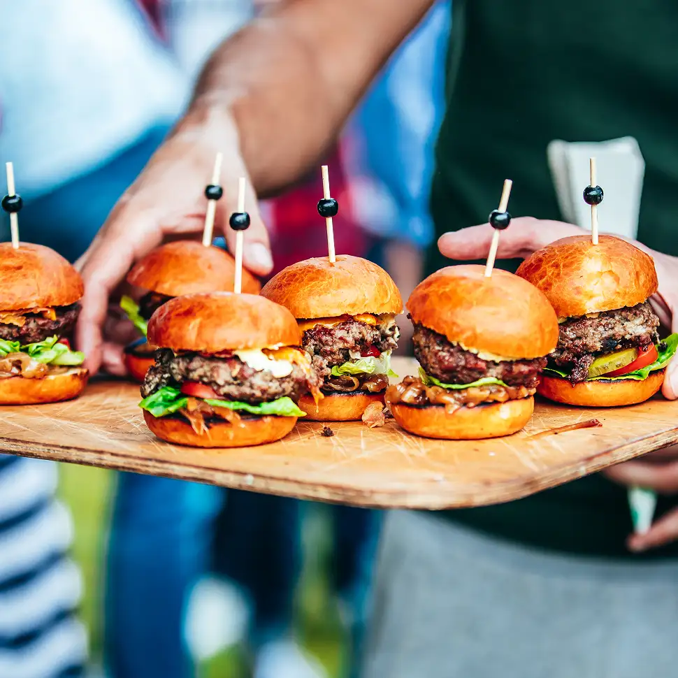 Person draagt een houten plank met zes kleine hamburgers met sla, tomaat en een satéprikker.