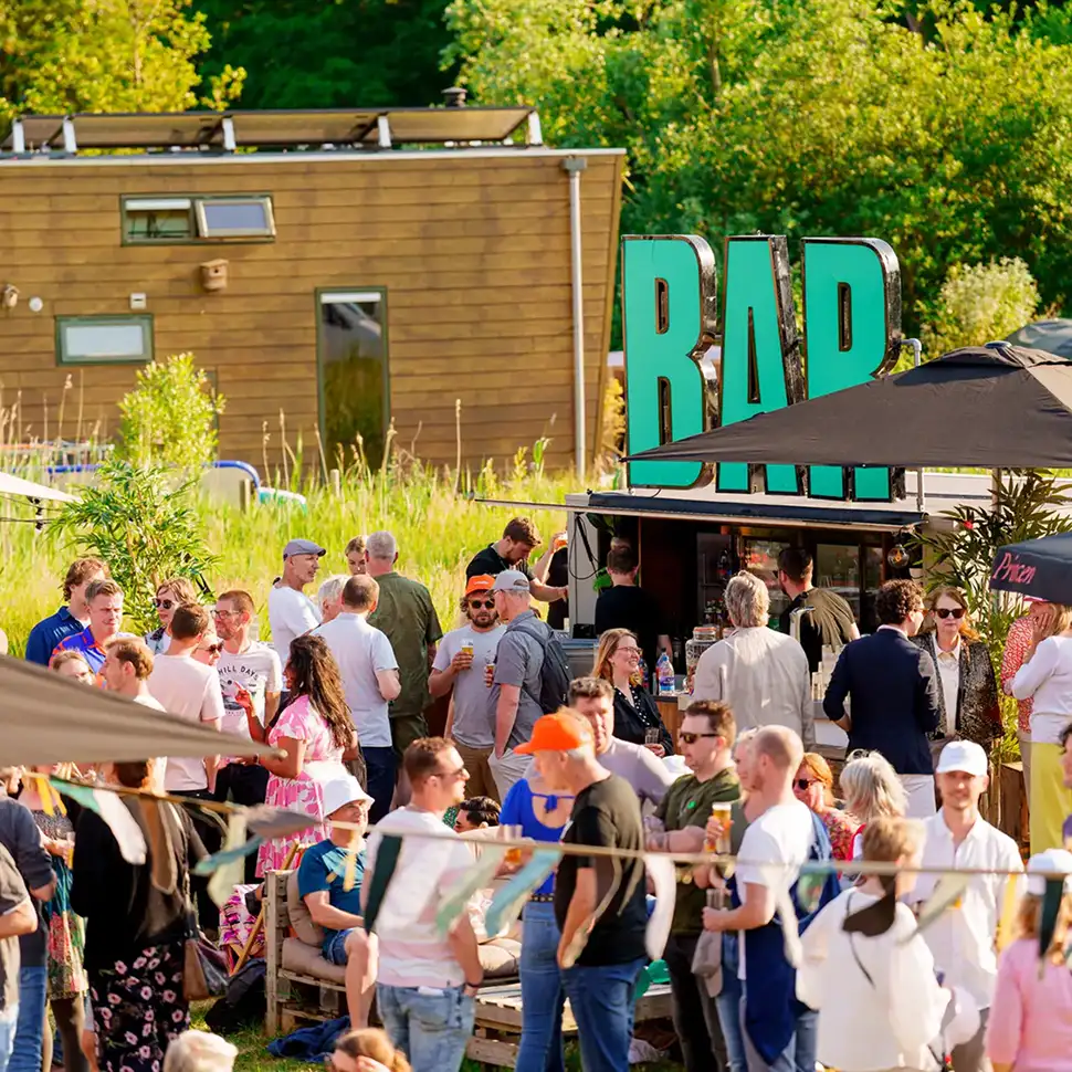 Mensen verzamelen zich bij een buitenbar met een groot bord 'BAR' tijdens een zonnige dag.