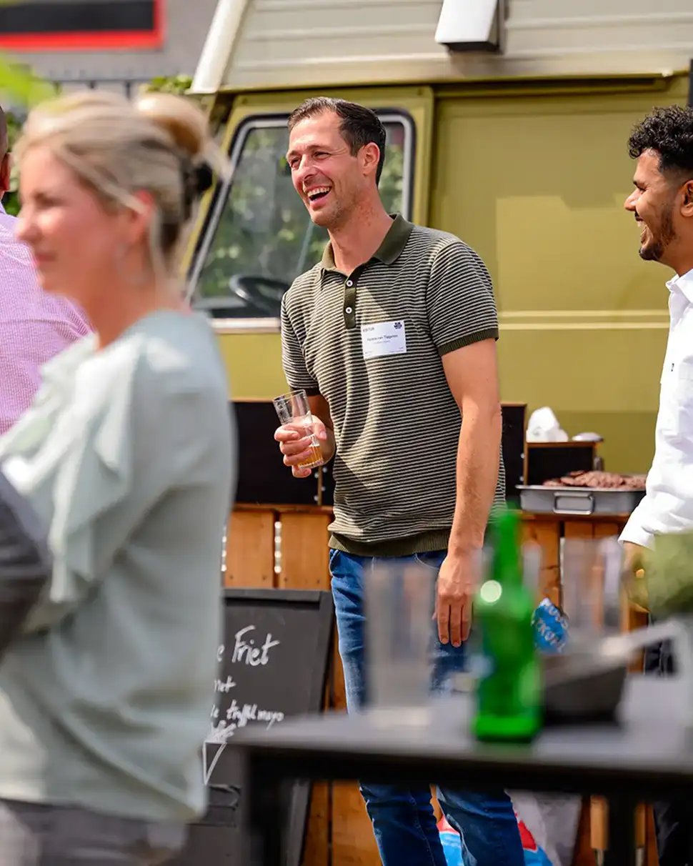 Mannen die lachend met elkaar praten bij een buitenbijeenkomst met een groene bestelwagen op de achtergrond.