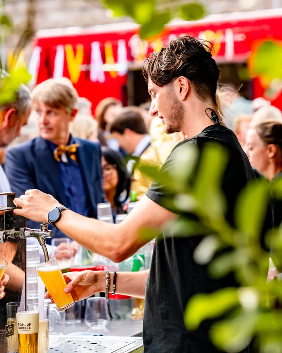 Man schenkt een glas bier in bij een drukke bar met mensen op de achtergrond.