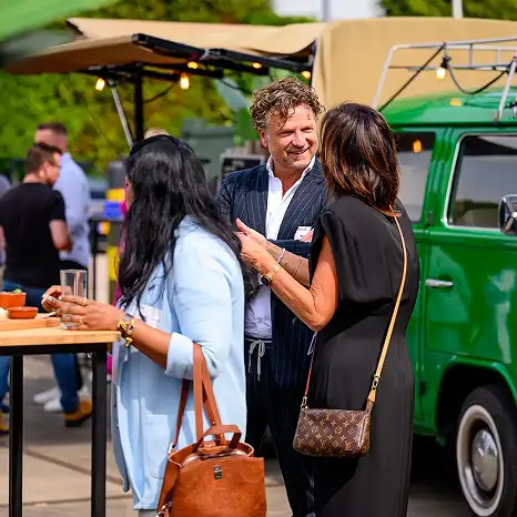 Drie volwassenen praten en lachen buiten bij een evenement naast een groene camper van Volkswagen.