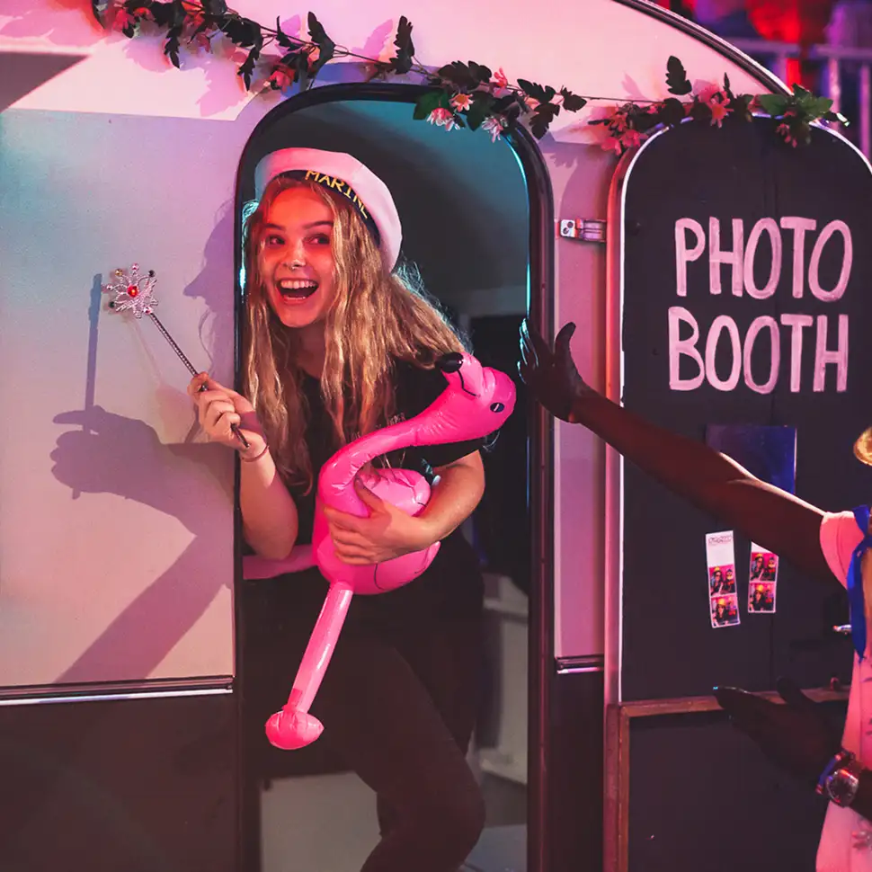 Vrolijke vrouw met een feesthoed en opblaasbare roze flamingo komt uit een fotohokje met bloemenversiering.