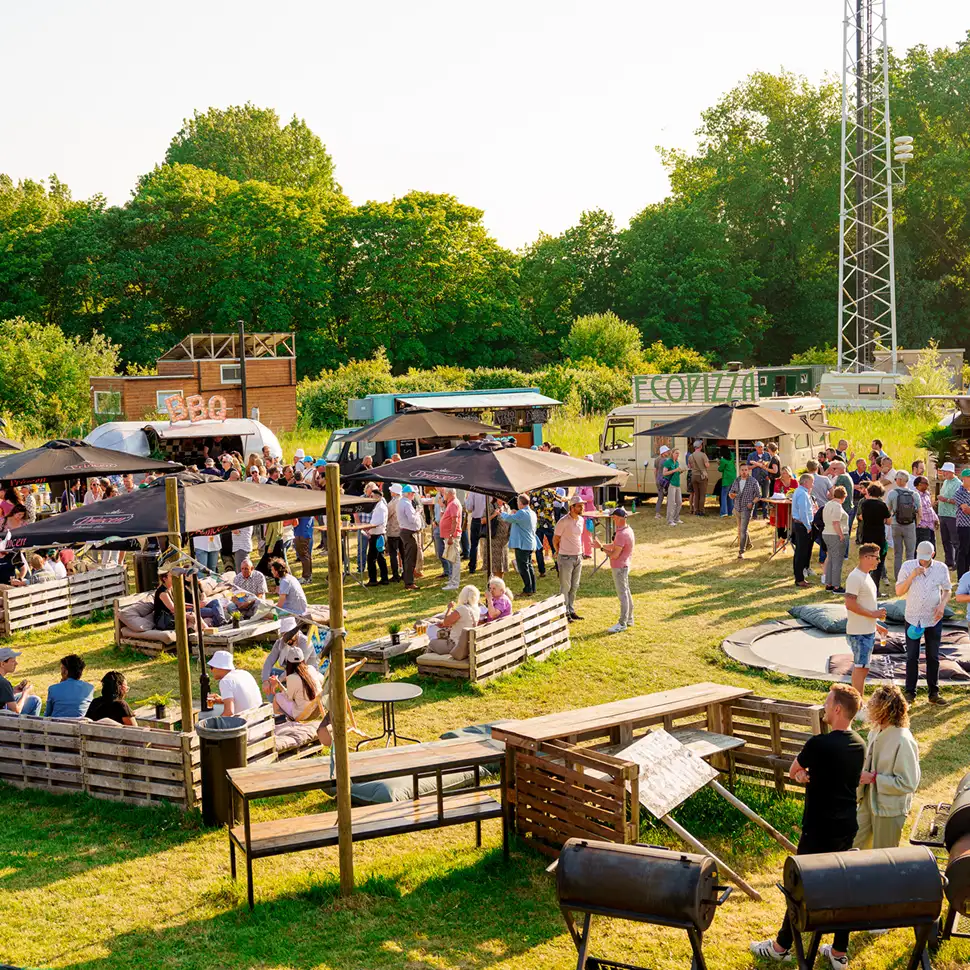 Buitenfeest met mensen die socializen rond foodtrucks, houten zitjes en parasols op een grasveld bij zonlicht.