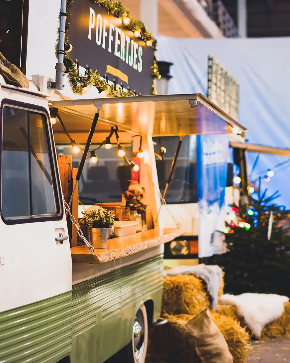 Foodtruck met het bord 'Poffertjes' versierd met kerstverlichting en houten balies, omringd door hooibalen en kerstbomen met lampjes.