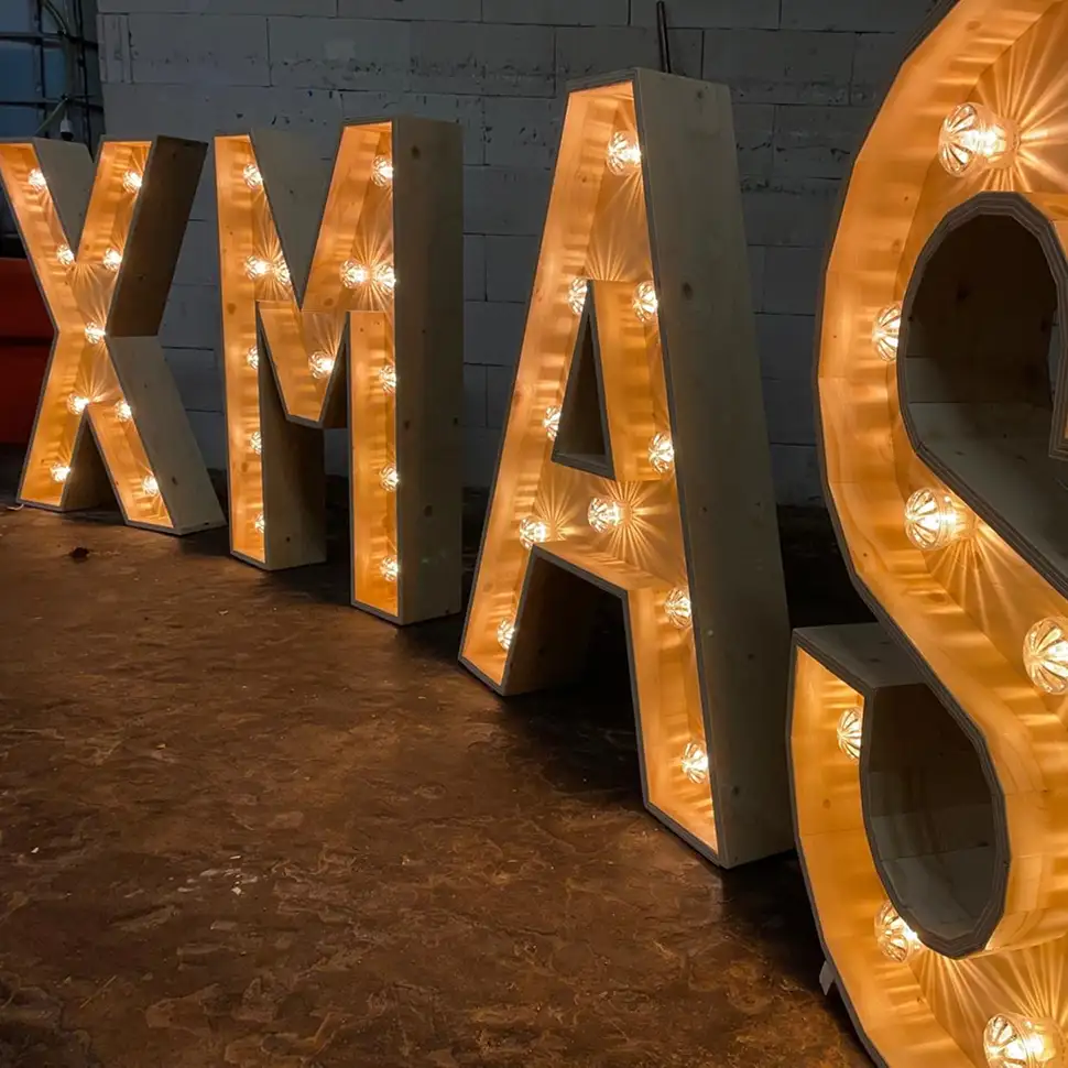 Grote houten letters met binnenin lichtbollen die het woord XMAS vormen, geplaatst op een donkere vloer tegen een bakstenen muur.