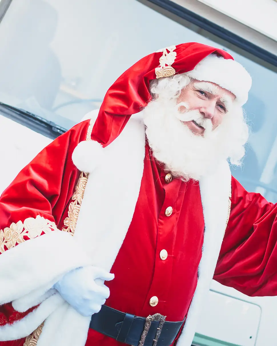Man in een traditionele kerstman outfit met rode jas, witte baard en muts die naar de camera kijkt.