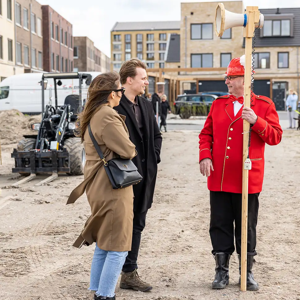 Twee mensen in jas en jas lopen op een bouwterrein en spreken met een man in een rood uniform en helm die een houten paal met een megafoon vasthoudt.