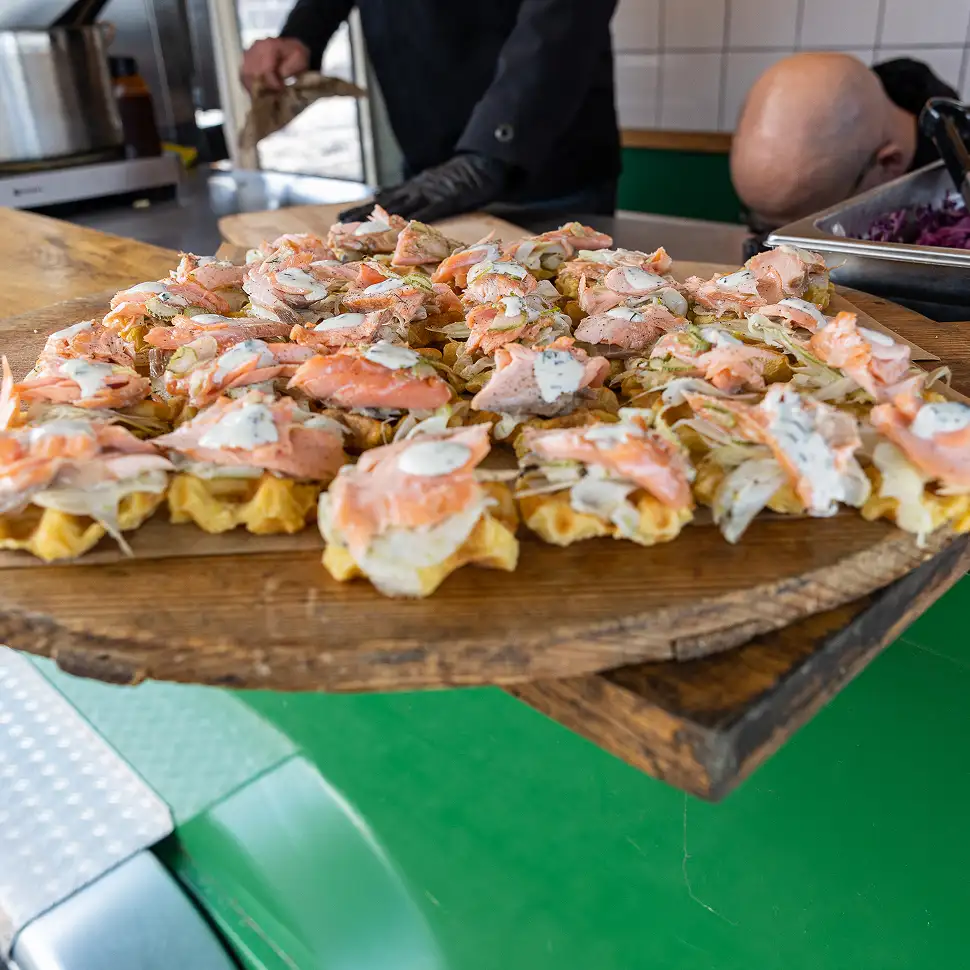 Houten plank met hapjes van wafels belegd met zalm, ui en een witte saus, gedragen door een persoon in een keuken.