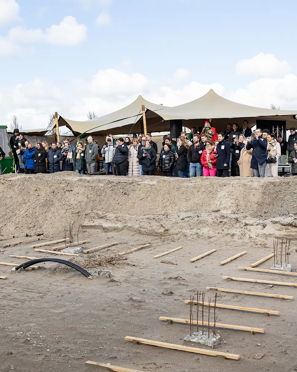 Groep mensen staat achter een bouwplaats met houten latten en betonnen fundamenten, onder een tent op een bewolkte dag.