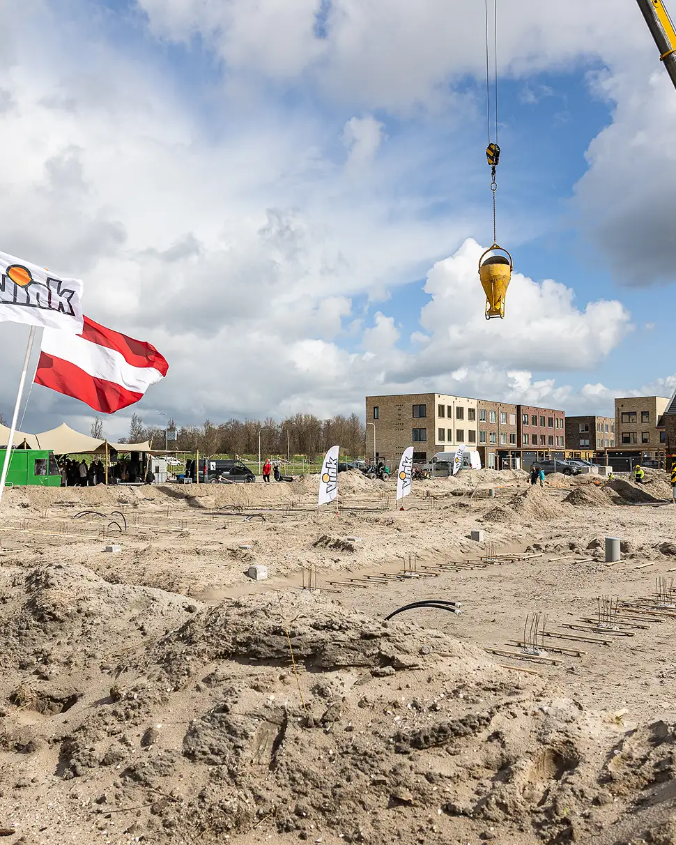 Bouwterrein met zandheuvels, hijskraan met geel betonemmertje in de lucht, vlaggen en moderne woningen op de achtergrond onder een bewolkte blauwe hemel.