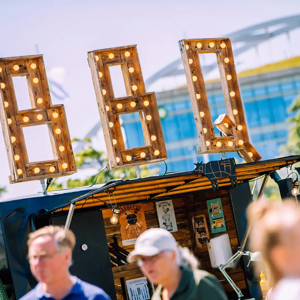 Verlichte houten letters BBQ op een foodtruck met wazige mensen op de voorgrond.