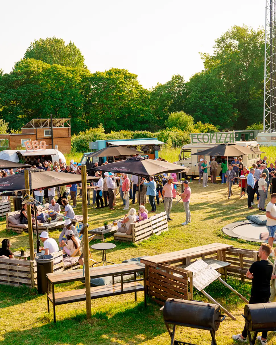 Buitenevenement met mensen die ontspannen rond houten zitjes en staan bij foodtrucks met BBQ- en ecopizza-borden, omringd door groen gras en bomen.