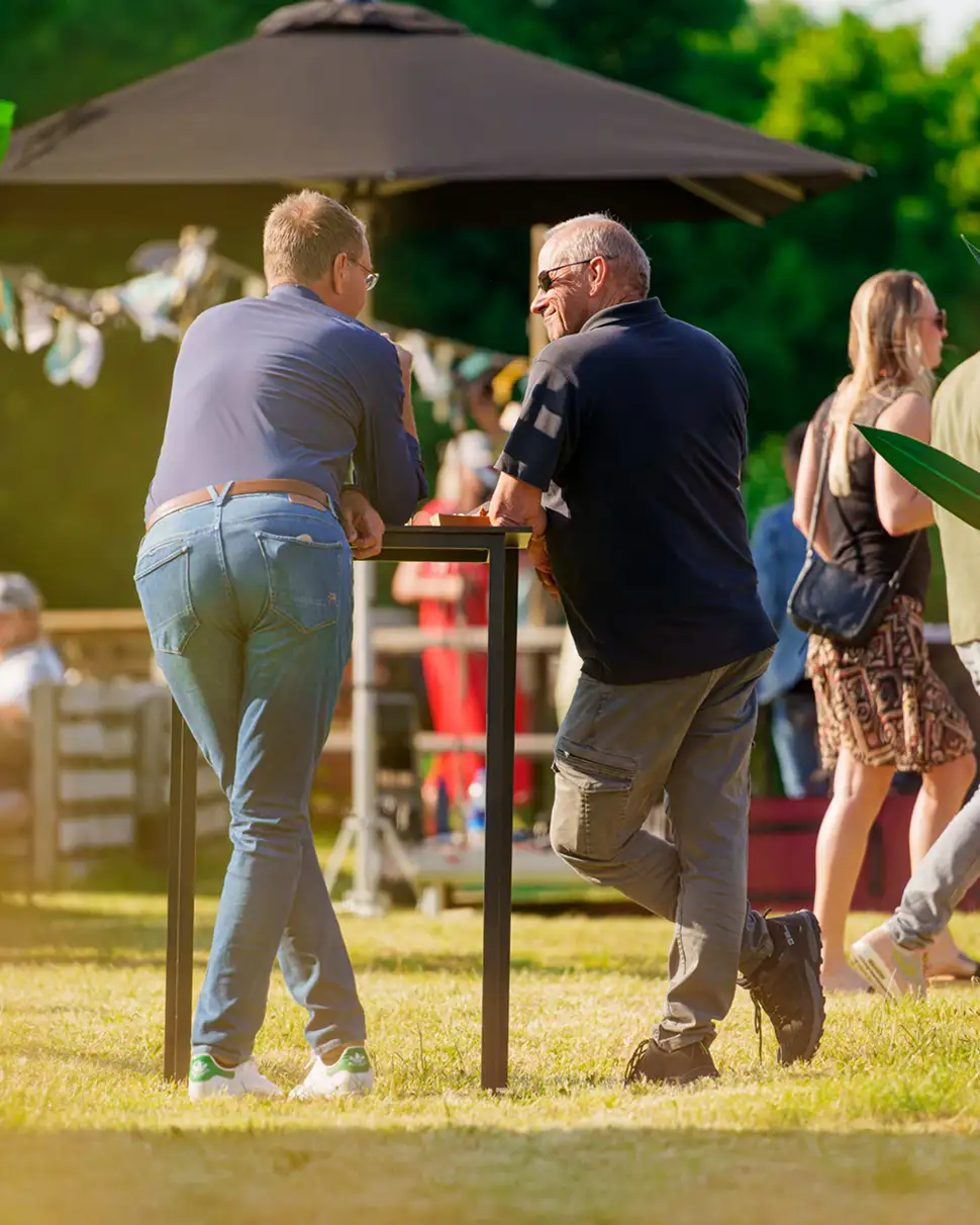 Twee mannen staan buiten bij een hoge tafel en praten tijdens een zonnige dag, op de achtergrond lopen andere mensen.
