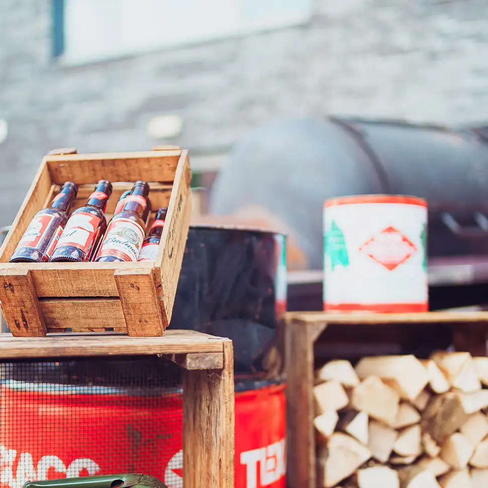 Houten krat met flesjes Budweiser bier naast gestapeld haardhout en metalen barbecue.