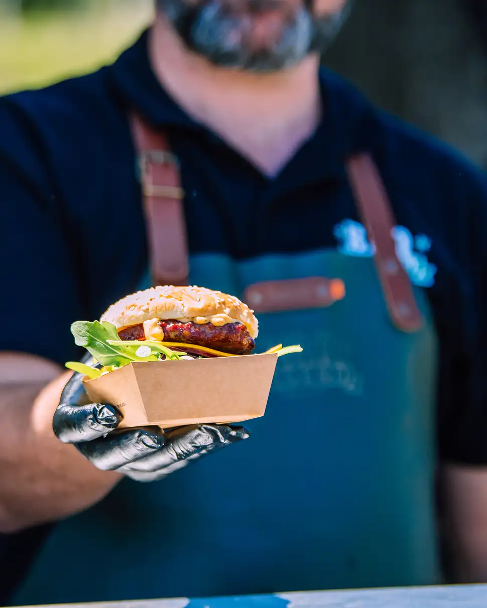 Man met zwarte handschoen houdt een hamburger met sla en saus in een kartonnen bakje.
