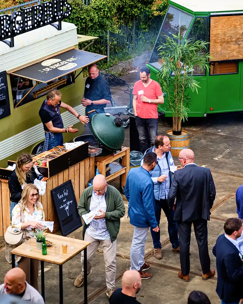 Drie lachende mannen achter een houten bar, gekleed in zwarte shirts met 'POP UP BORREL' tekst, onder rood licht.