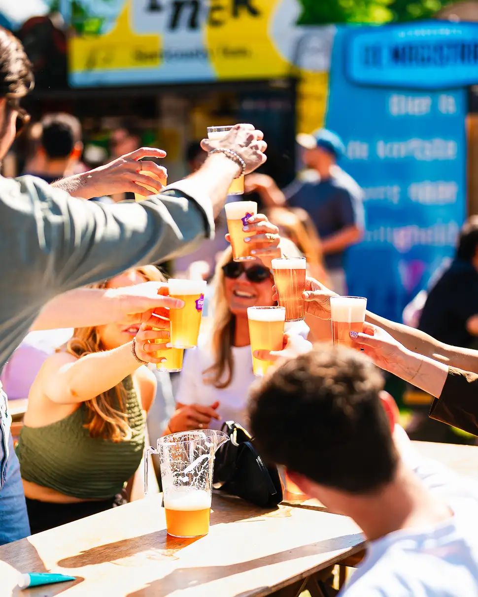 Man met zonnebril schenkt een glas bier in bij een buitenbar met groot groen bord 'BAR' op de achtergrond.