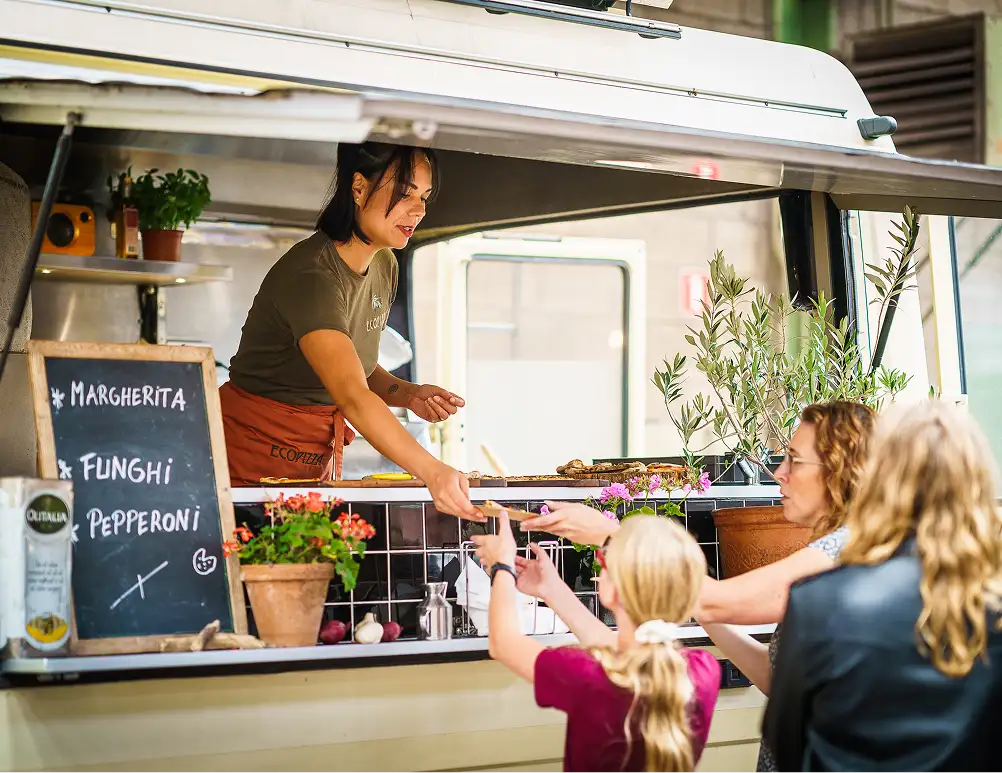 Vrouw serveert pizza van een foodtruck aan een vrouw en een kind in de rij, met een bord met pizzasoorten en planten op de toonbank.