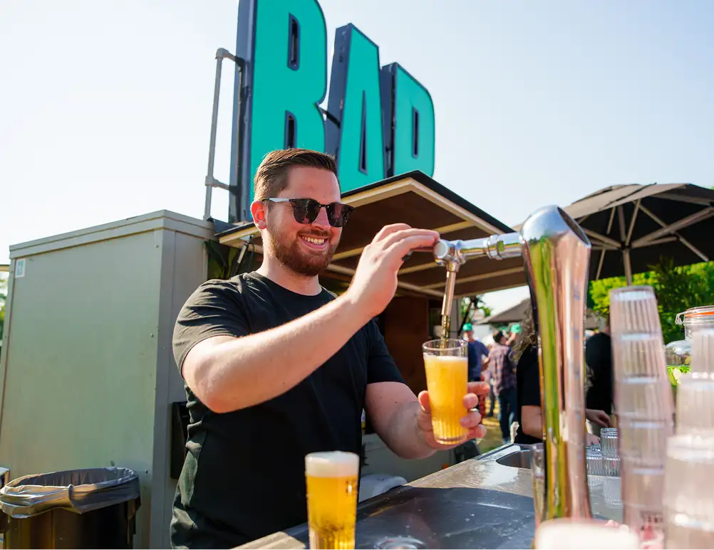 Man met zonnebril glimlacht terwijl hij een glas bier tapt aan een bar in de open lucht.