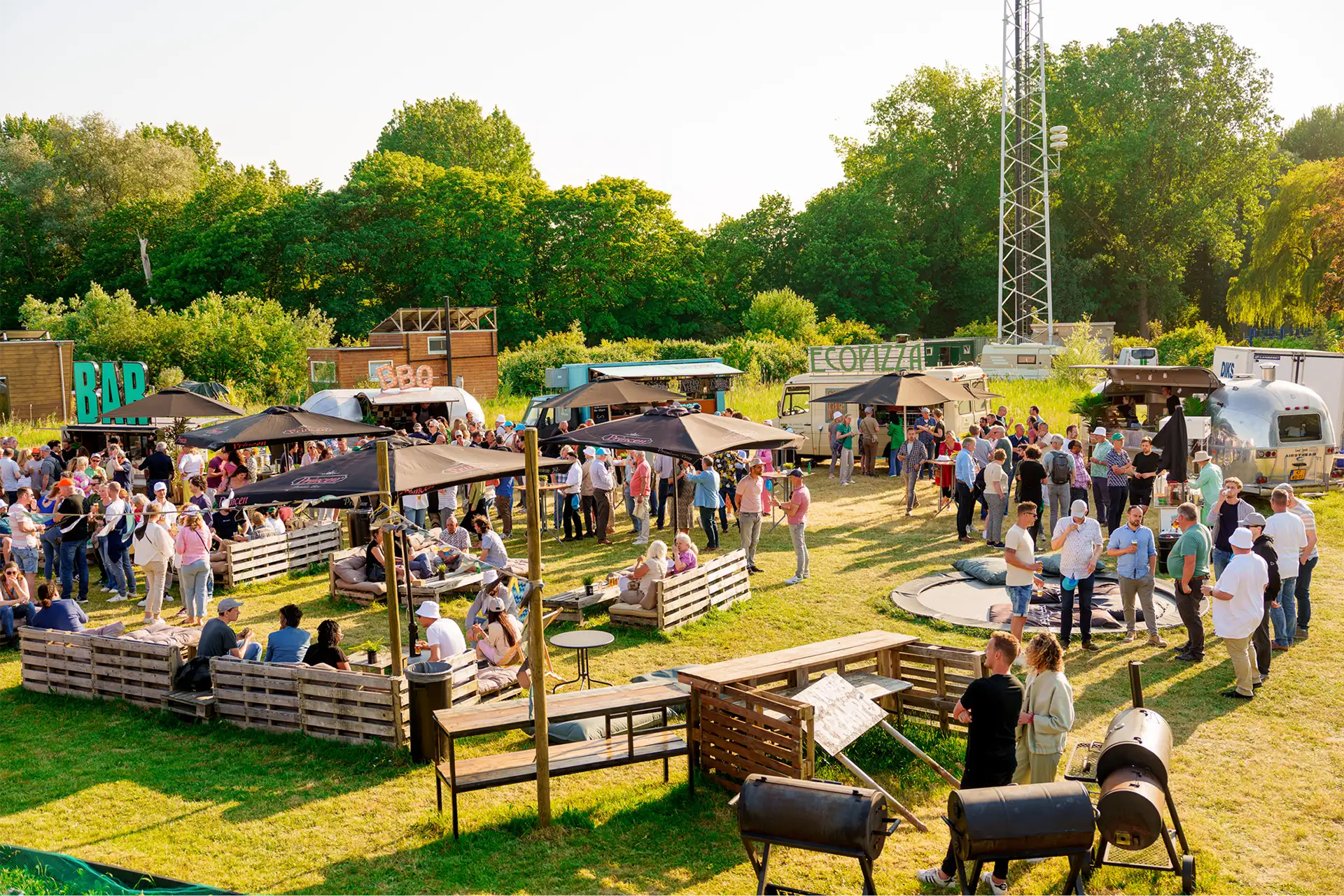 Mensen genieten van een zomerse borrel in een buitenruimte met bar, BBQ en foodtrucks, omgeven door groen.