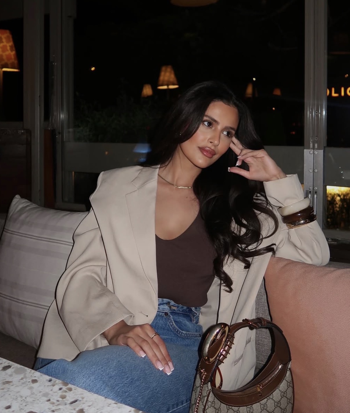 Woman with long dark hair wearing a beige blazer, brown top, and blue jeans sitting on a couch with a designer handbag nearby.