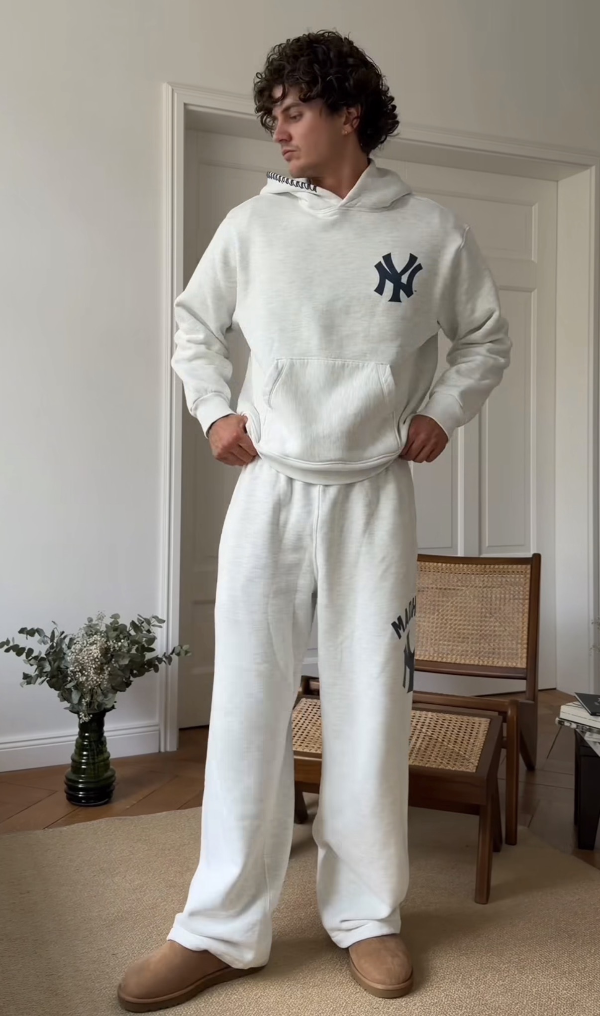 Man wearing light gray New York Yankees hoodie and matching sweatpants with tan slippers standing indoors.