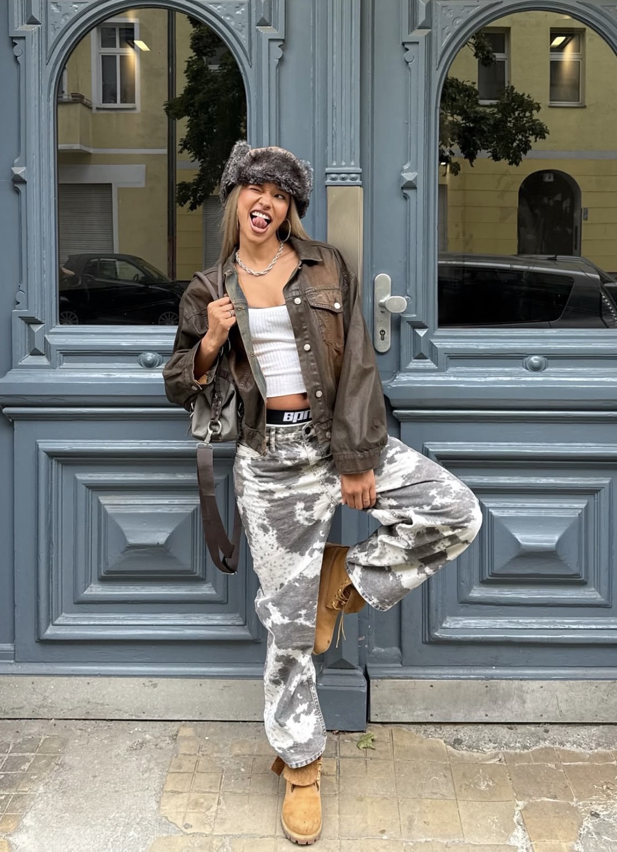 Young woman in a furry hat, cropped jacket, white tank top, and tie-dye pants posing playfully with tongue out and one leg raised.
