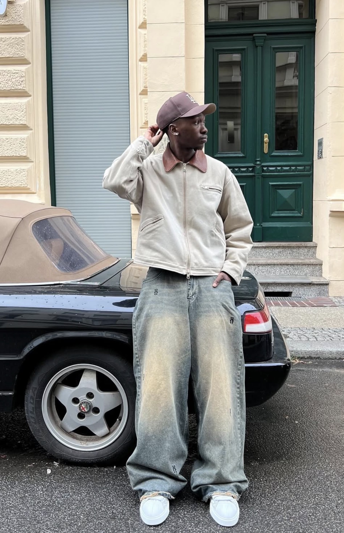 Man wearing a beige jacket, baggy faded jeans, white sneakers, and a purple cap standing in front of a black convertible car with a green door in the background.