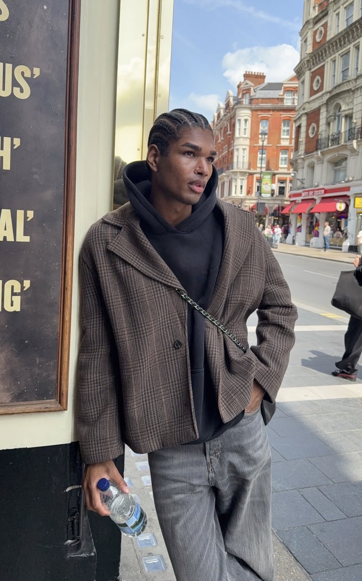 Young person with braided hair wearing a plaid jacket and black hoodie, leaning against a wall holding a water bottle on a city street.