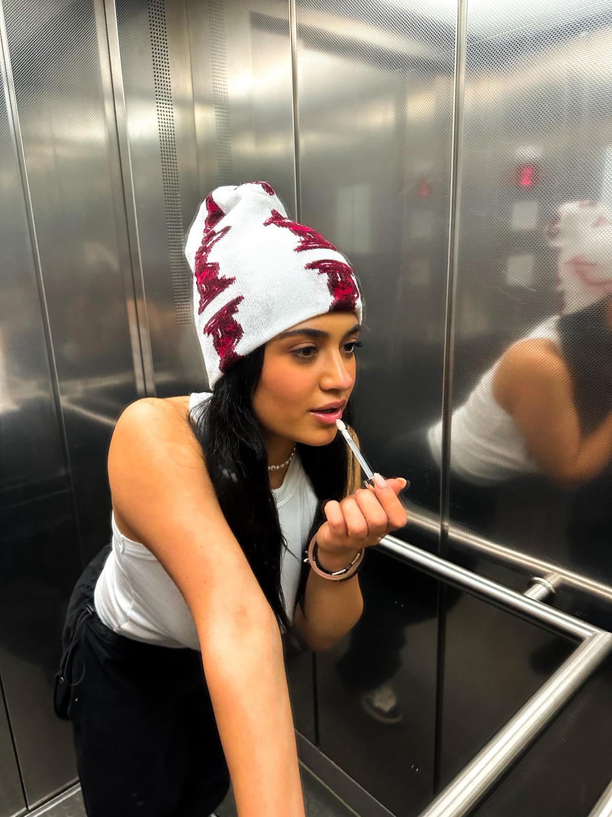 Young woman wearing a white and red knit hat applying lip gloss in a metallic elevator.