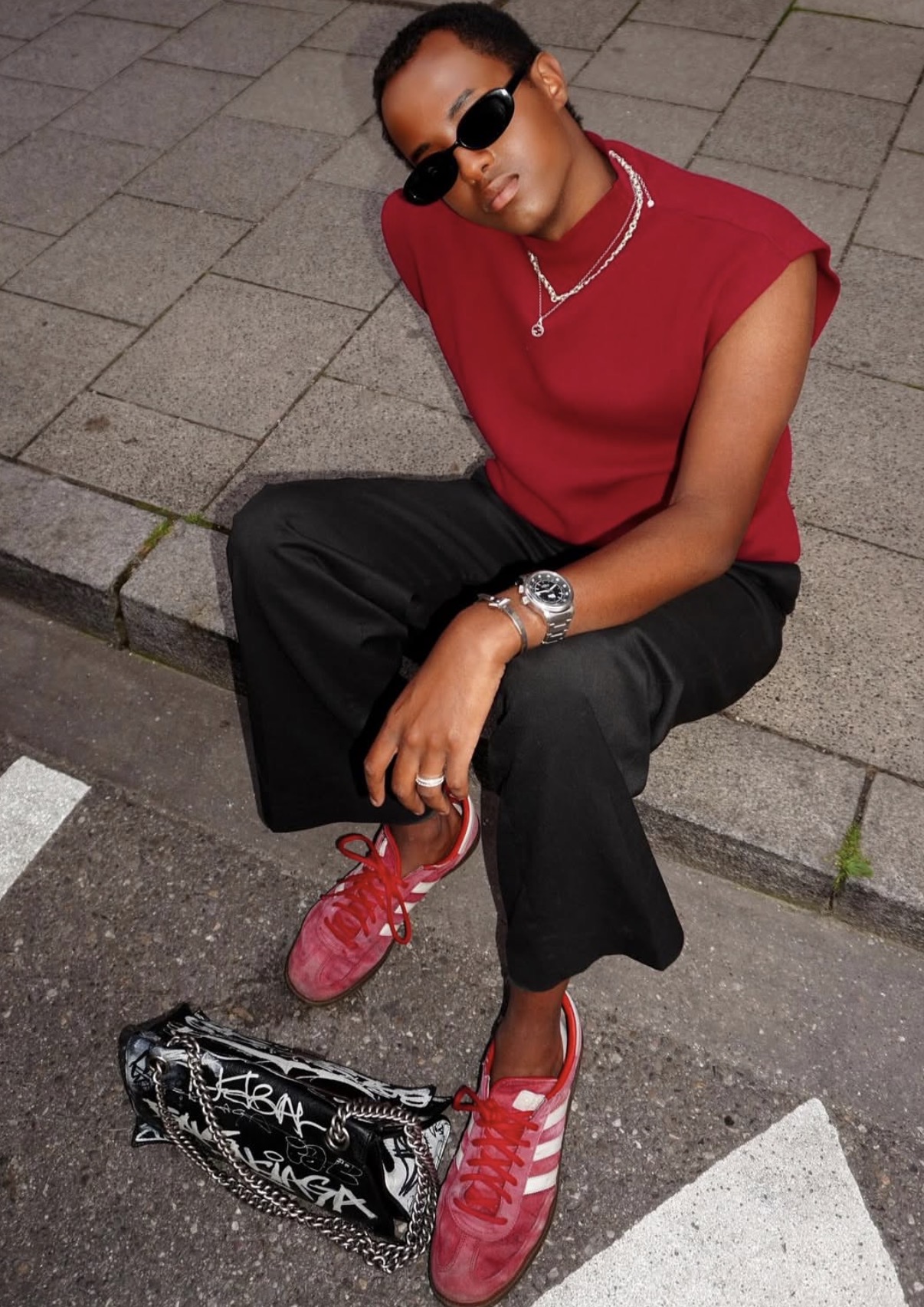 Person wearing sunglasses, a red sleeveless top, black wide-leg pants, and red Adidas sneakers sitting on pavement with a black graffiti-patterned bag nearby.