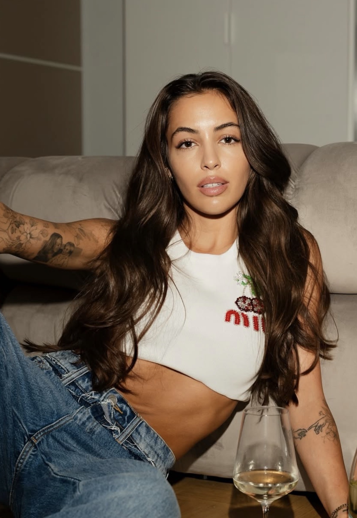 Young woman with long dark hair reclining on a beige sofa, wearing a white crop top and blue jeans, with tattoos on both arms and a glass of white wine in front.